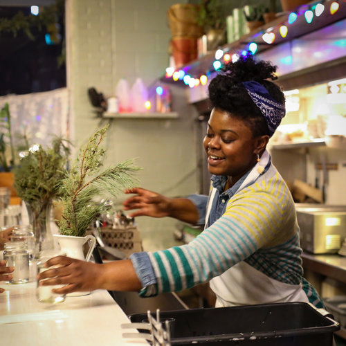 Kezia behind a white bar, with string lights on the wall behind, smiling and serving water