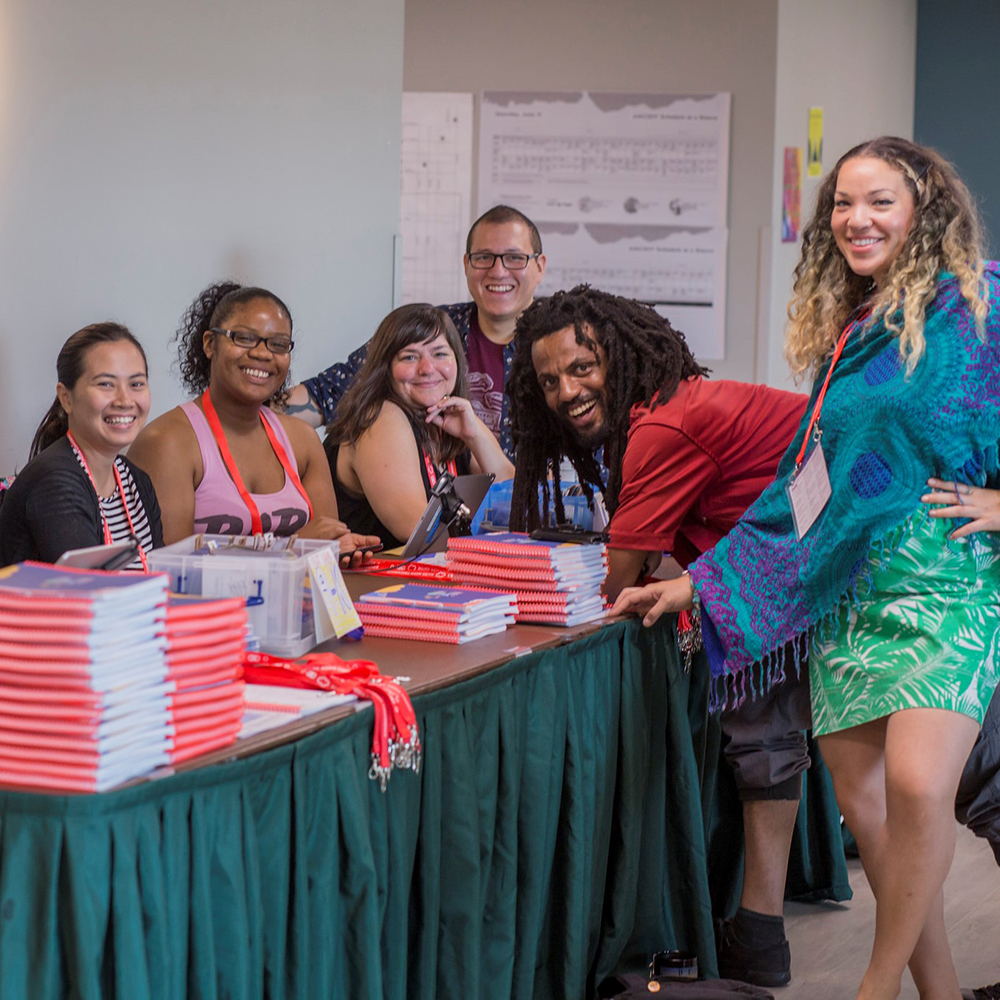 group of 6 volunteers sitting and standing around the AMC registration table, which is stacked with AMC guides and lanyards.