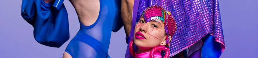 Person's face with bright makeup and head adornment, with two people in the background wearing a bodysuit and a bright purple cape