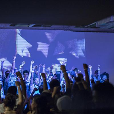 Photo from the Allied Media Conference, with people on stage and in the audience standing with fists raised in solidarity.
